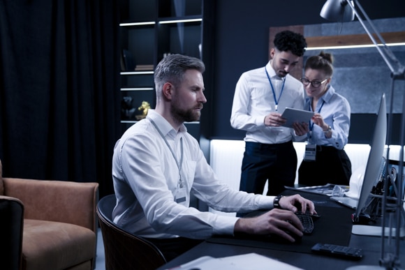 Mitarbeiter am Schreibtisch optimiert die IT Sicherheit, im Hintergrund unterhalten sich zwei Kollegen.